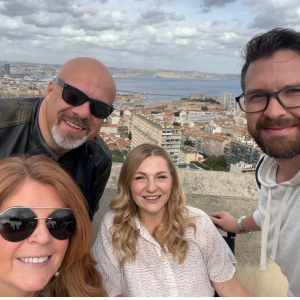 Group of four smiling individuals posing for a selfie with a scenic city view in the background, highlighting camaraderie and support for community initiatives.