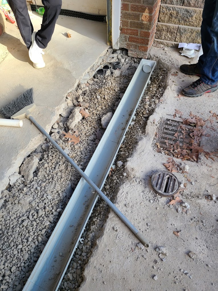 A construction site shows a partially installed drain and trench in front of a building with visible gravel. Two people stand nearby with tools.