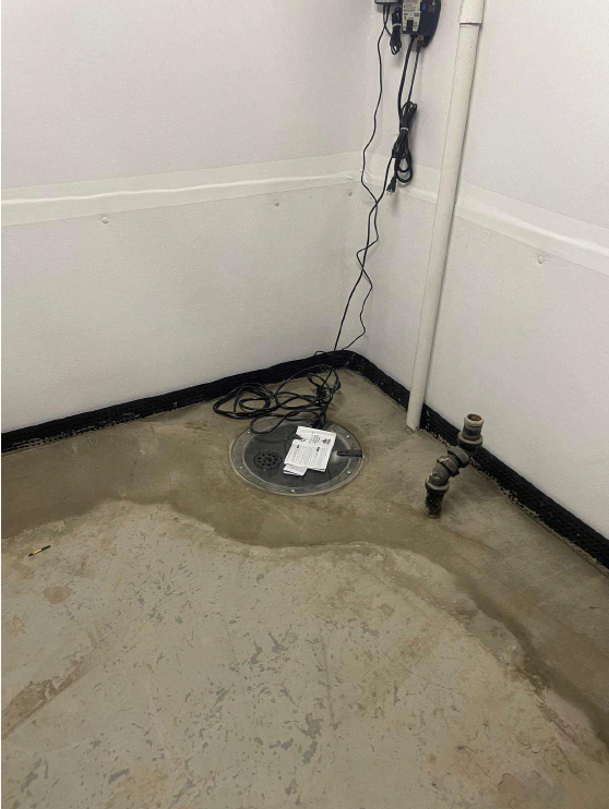 A plain concrete basement corner with white walls is shown. A drain on the floor is surrounded by wires and pipes. A single paper is on the floor.