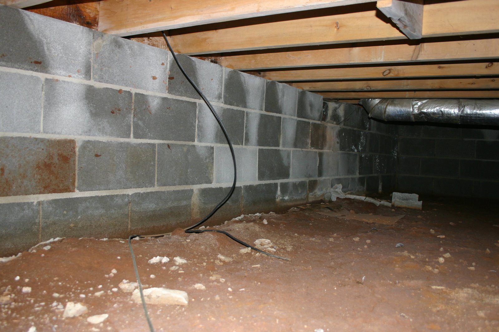 A dimly lit crawl space with a dirt floor and cinder block walls. Exposed wooden beams support the ceiling, and there are wires and ducts visible.