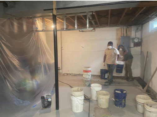 Two people in masks work in an unfinished basement. They carry buckets near plastic sheeting, surrounded by exposed beams and a dim light. The scene feels industrious.