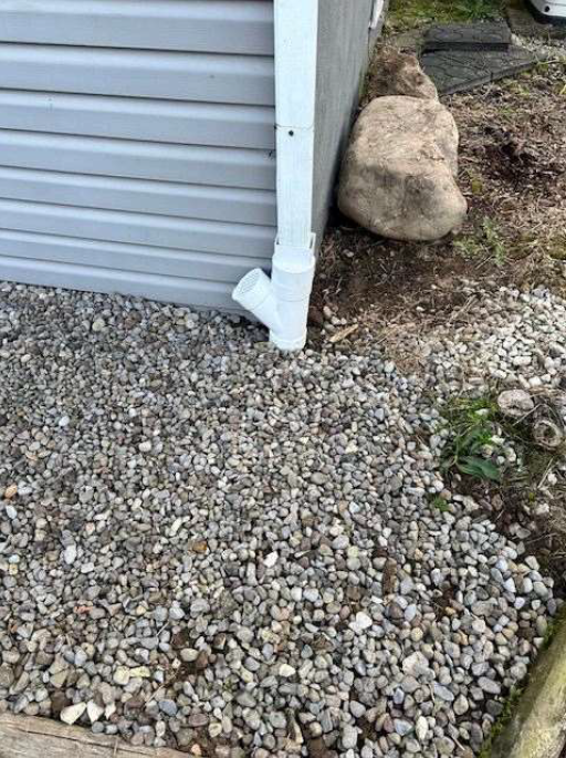 A white downspout on the corner of a gray building drains onto gravel. A large rock and some grass are visible nearby, hinting at an outdoor setting.
